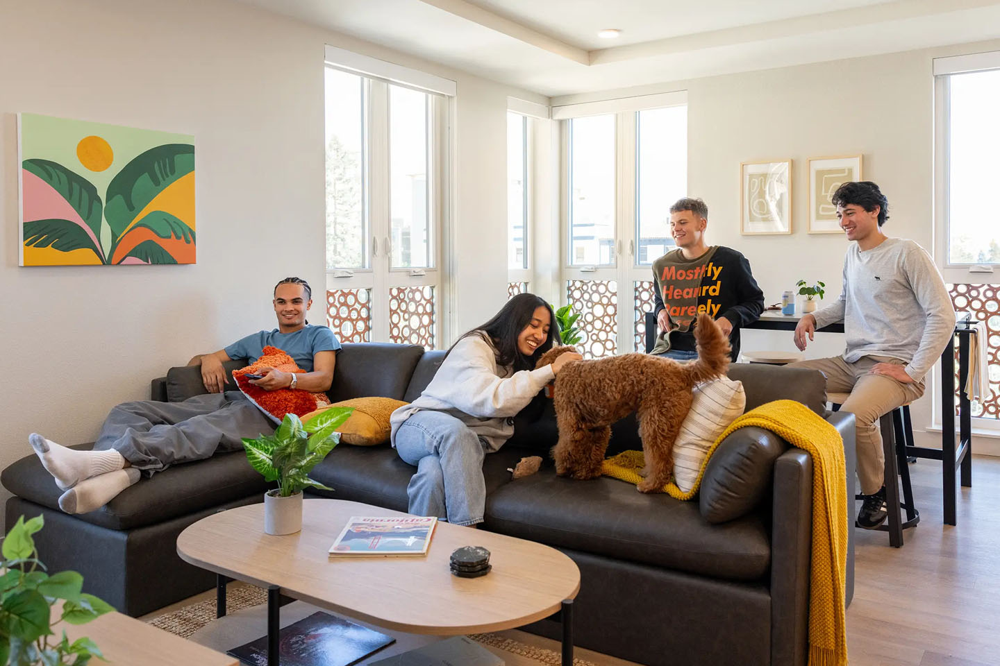 Friends in a living room space, two on couch two sitting at the table playing with cute brown dog.